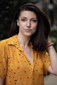 Headshot of Laura Davey; they have dark hair and are wearing a yellow shirt with polka dots. They stare directly into the camera. Ambient image