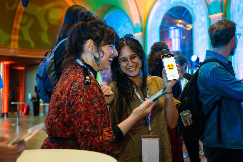 Two Attendees At The Conference Smile And Show Each Other Emojis On Their Phones